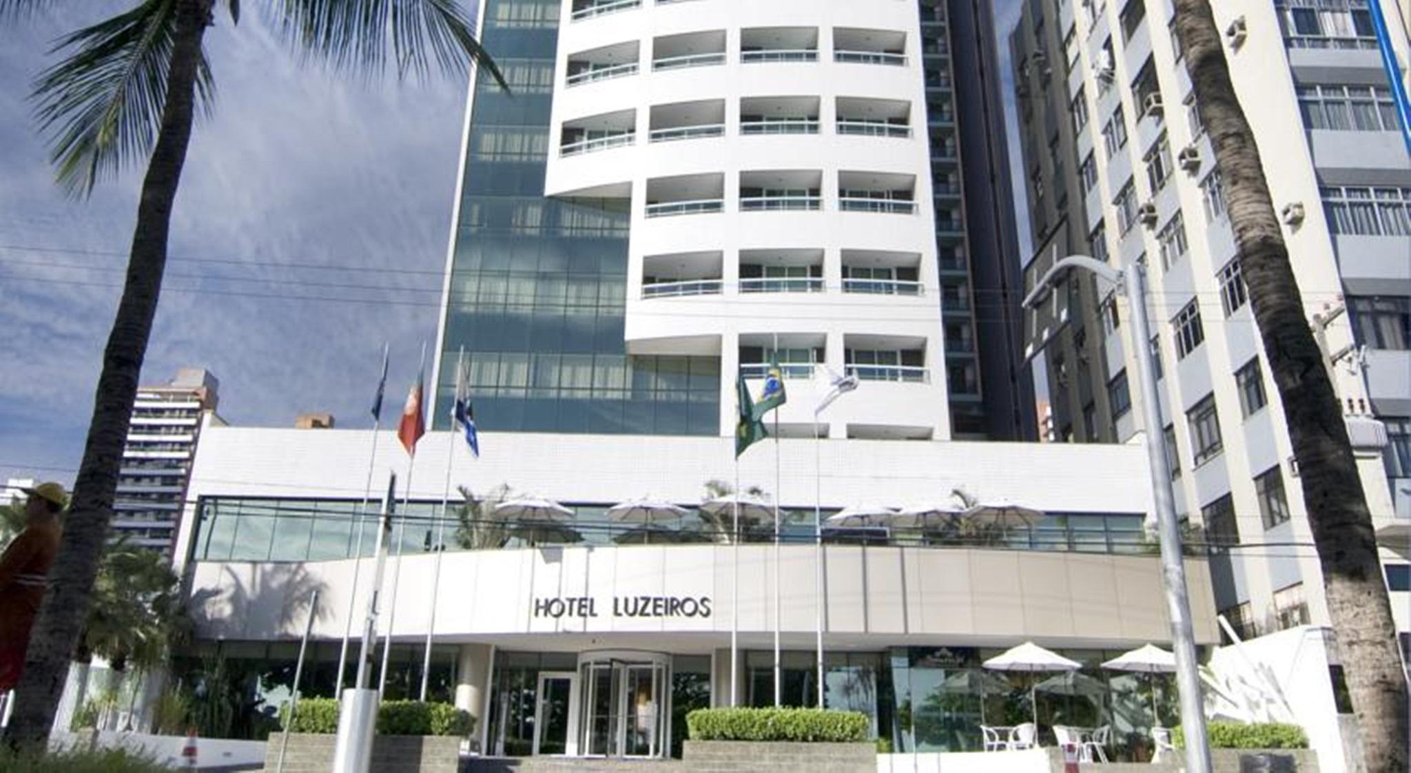 Hotel Luzeiros Fortaleza 외부 사진 The photo shows the facade of a modern hotel named "Hotel Luzieros." The building features a sleek design with a combination of glass and concrete elements. Several flags are displayed in front, suggesting an international appeal. The entrance area i