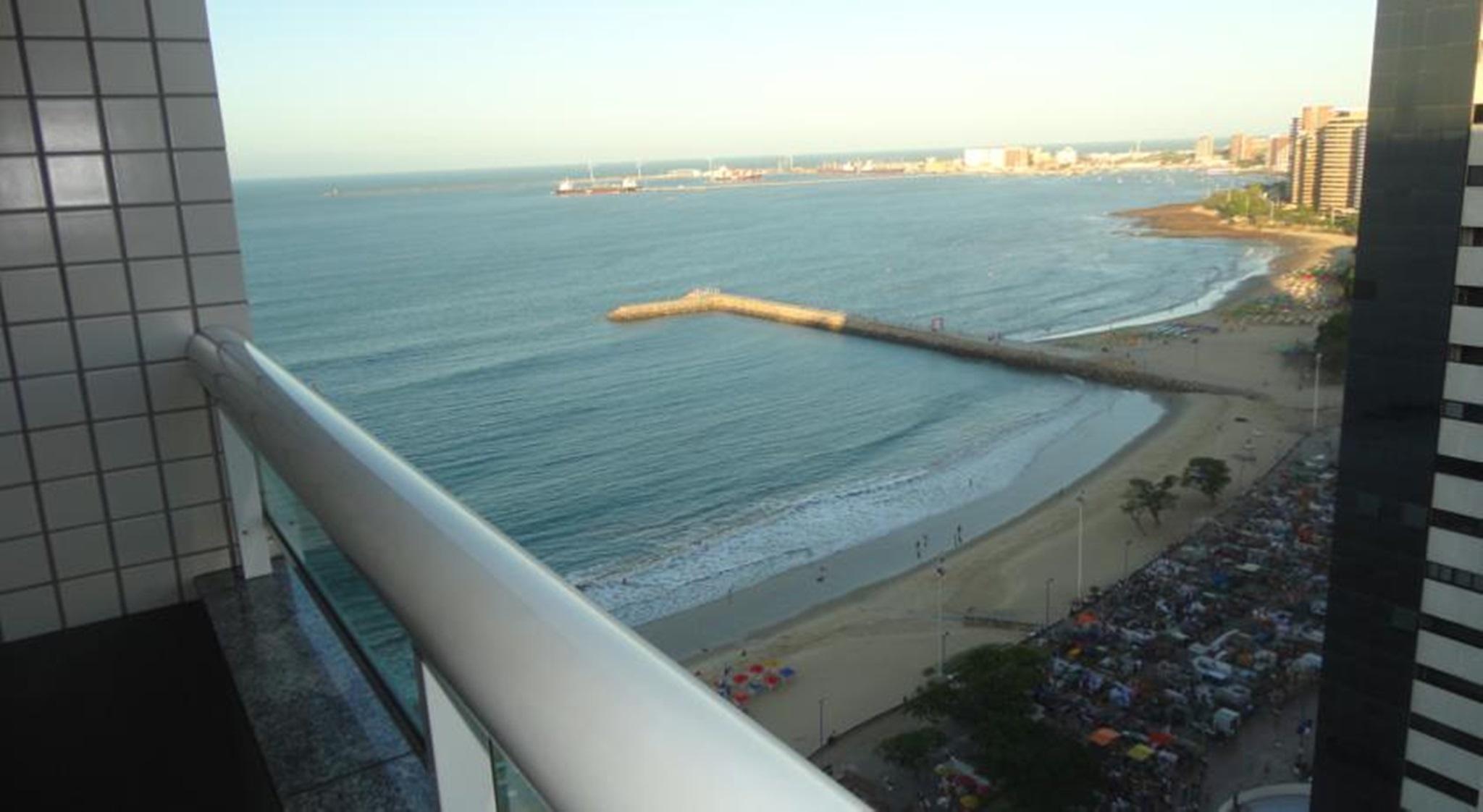 Hotel Luzeiros Fortaleza 객실 사진 The photo depicts a coastal scene taken from a high point, possibly a balcony. In the foreground, there is a white railing, and you can see the shoreline with gentle waves lapping at the sand. There appears to be a small pier extending into the water
