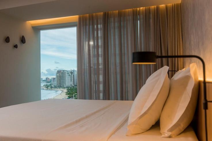 Hotel Luzeiros Fortaleza 외부 사진 The photo shows a modern and stylish bedroom. The bed is adorned with two large, fluffy pillows, and the bedding appears to be neatly arranged. A warm light fixture is mounted on the wall beside the bed, adding a cozy ambiance. There are large window