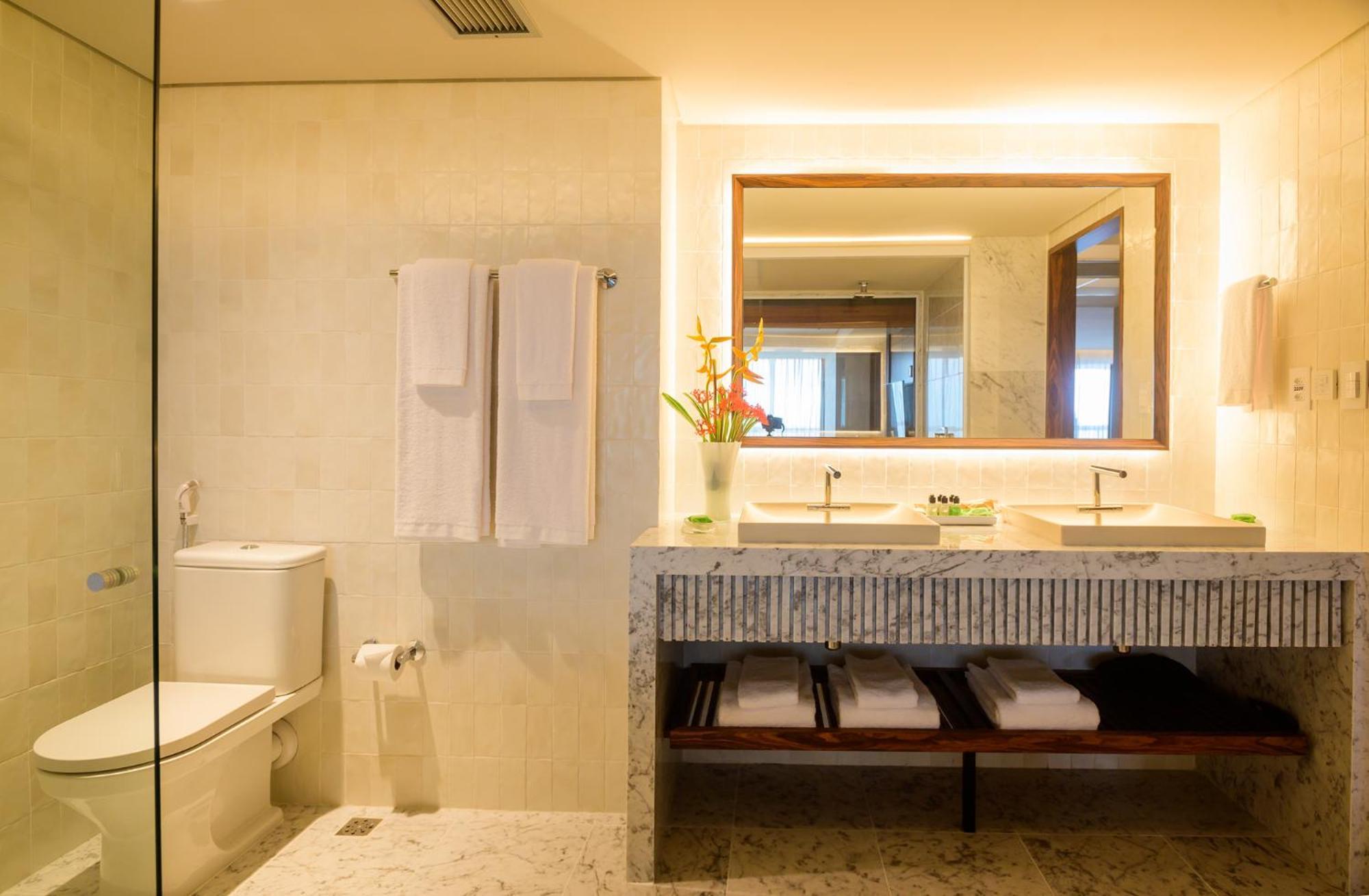 Hotel Luzeiros Fortaleza 외부 사진 The photo shows a modern bathroom. It features a glass shower enclosure, a white toilet, and a double sink vanity made of stone. Above the sink, there's a large mirror with warm lighting around it. Hand towels hang neatly on a rack, and there are som