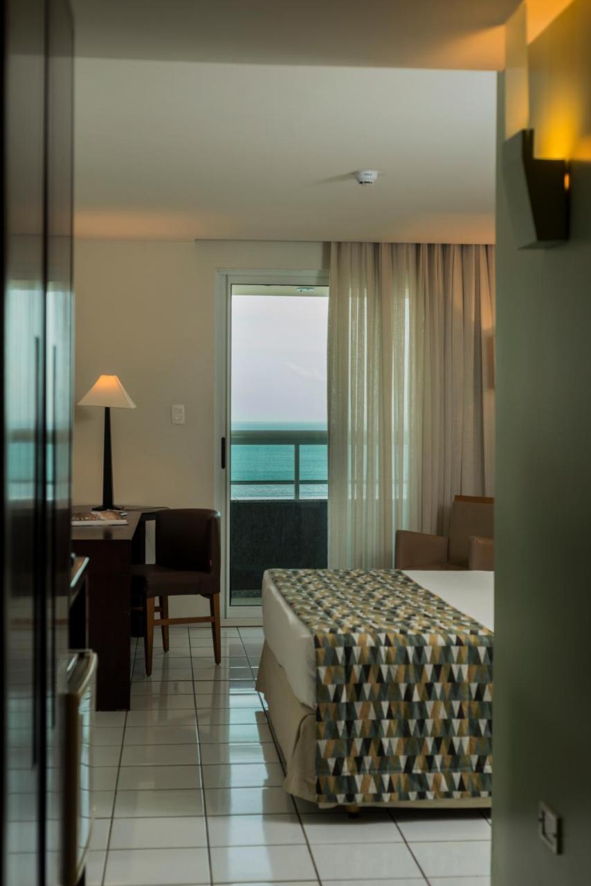 Hotel Luzeiros Fortaleza 외부 사진 The photo showcases a hotel room with a balcony view. It features a bed with a patterned blanket, a bedside table, and a lamp. There is a small wooden desk with a chair in the corner. Sheer curtains frame a sliding glass door that opens to the balcon